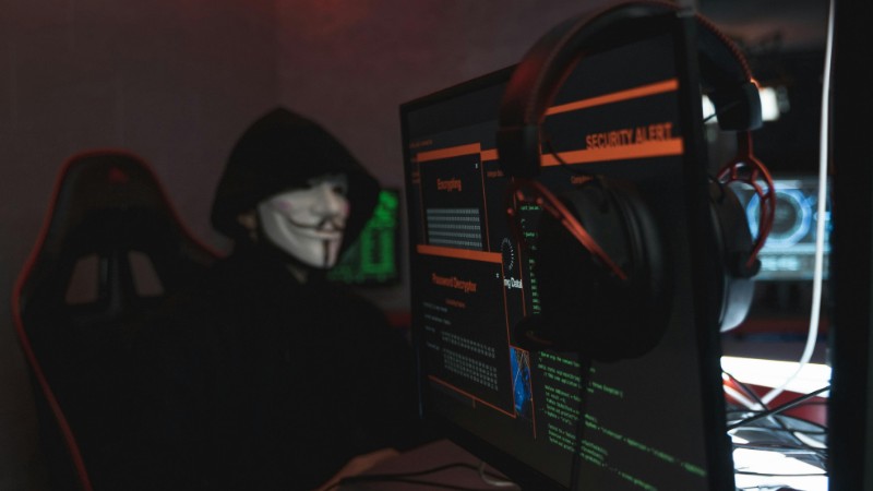 A person wearing a Guy Fawkes mask engaged in hacking activities on a computer in a dimly lit room., tags: cyberangriff ein - pexel